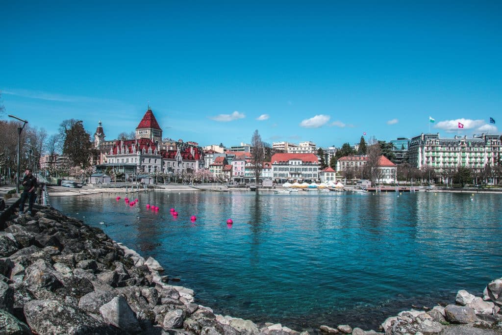 Lake in the city of Lausanne, Switzerland. The city is charming with its beautiful architecture. A pile of rocks can be seen in the foreground of the photo. A man is standing on one of the rocks. The sky is blue and clear.