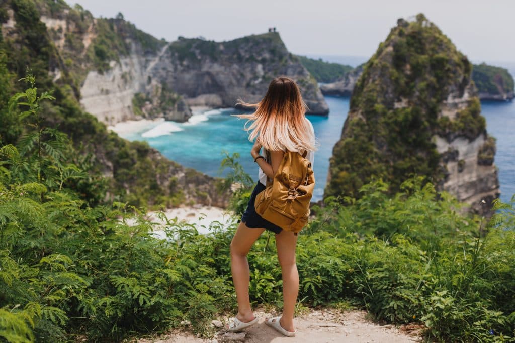 A young woman is hiking on a mountain on a paradise island, carrying a backpack. She is wearing a white tank top, navy blue shorts and white sandals. She has her back to the camera and is admiring the view from the vantage point.