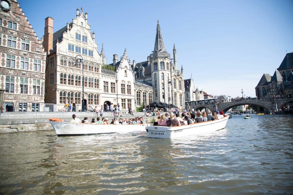 Groups of tourists are taking a boat trip on the canal in the city of Ghent, Belgium. They are admiring the typical Belgian buildings lining the canal.