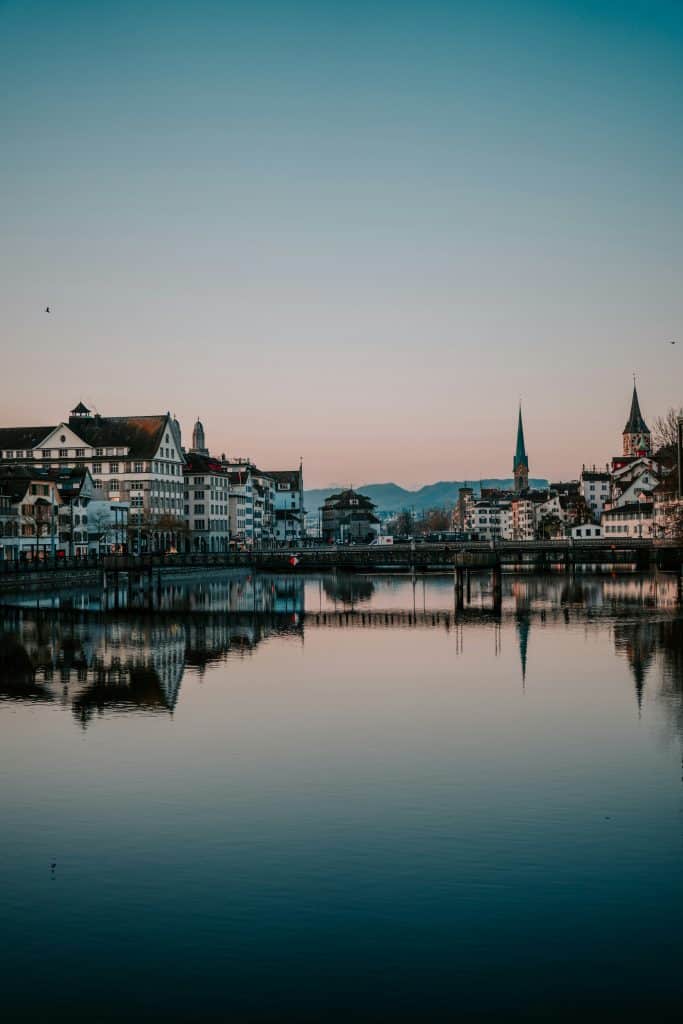 Portrait photo of the city of Zurich in Switzerland. The buildings are reflected in the water of a lake. The photo was taken in the early evening as the sun was setting.
