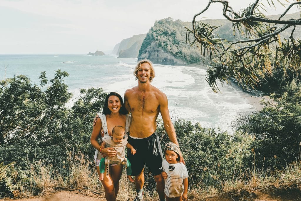 Family photo of a couple and their two children during a hike in a tropical destination. They are standing at the top of a hill, with a view behind them of an empty beach and cliffs.