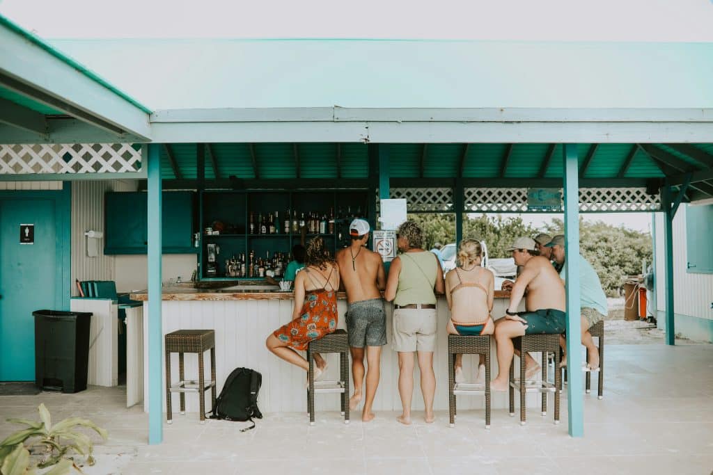 A family gathered around a bar in a tropical setting, some people are seated and others are standing. They are all dressed in swimwear or beachwear and have their backs to the camera. They are chatting among themselves.