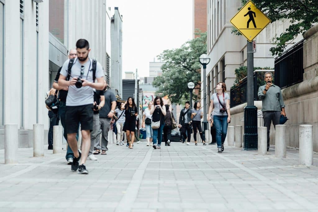 People walking on the concrete street in the middle of the day. Most of them are wearing t-shirts and looking at their phones as they walk.