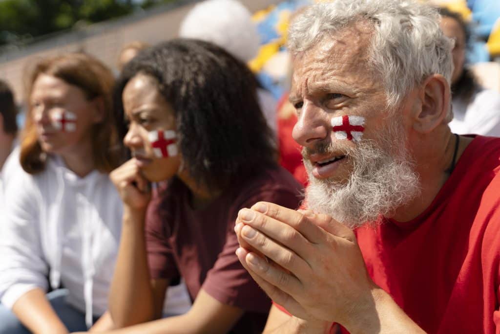 People sitting in the stands of a stadium display Swiss flags on their cheeks in support of their favourite team. They are focused on the match, and the elderly man in the foreground joins his hands in prayer, hoping to see his team score points.