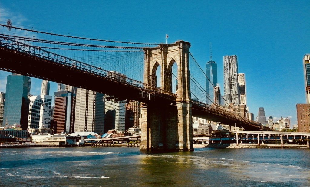 Photo of the Brooklyn Bridge in New York, taken on a very sunny day. The skyscrapers stand out in the background. The sea is slightly choppy.