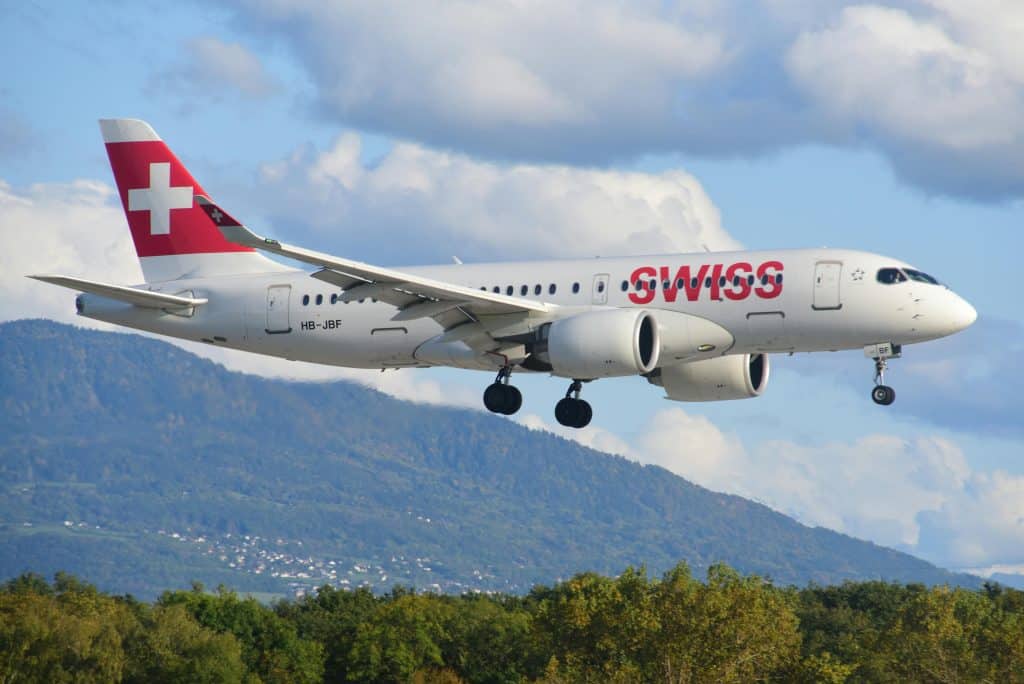 A Swiss Airlines plane is preparing to land at Geneva Airport. The sky is clear and mountains and forests can be seen in the background of the photo.
