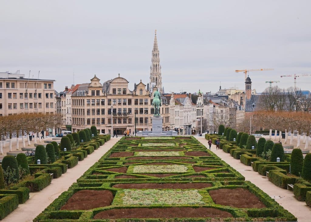 Photo of a beautiful garden in the capital city of Brussels, Belgium. The hedges and shrubs are neatly trimmed. A statue stands at one end of the garden. The sky is grey. The buildings around the square are well maintained.
