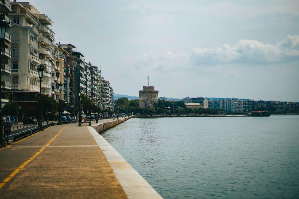 Photo of a body of water in Thessaloniki, Greece. A few people are walking along the quay. A tower with a flag can be seen in the background.
