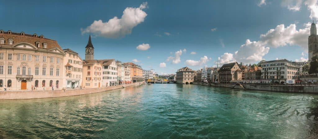 View of the river in the city of Zurich, Switzerland. Beige buildings line both sides of the riverbank. A bridge can be seen at the end of the photo.