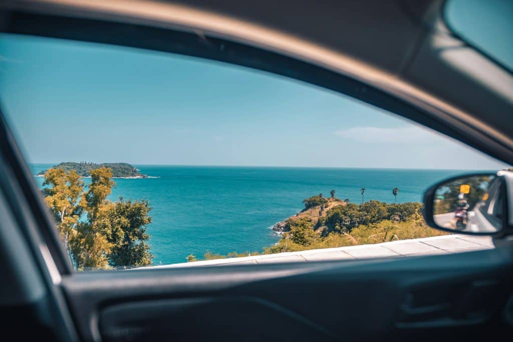 Photo taken from inside a car in the driver's seat, looking outwards with the window rolled down. The view shows a picturesque coastal landscape and a lone island in the middle of the sea. In the rear-view mirror, two people on a motorbike can be seen preparing to overtake.