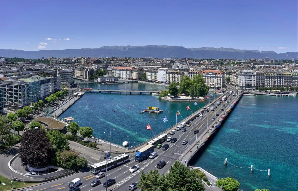 Aerial photo of the city of Geneva in Switzerland. The photo overlooks a busy bridge in the city that connects different neighbourhoods. Mountains can be seen on the horizon.
