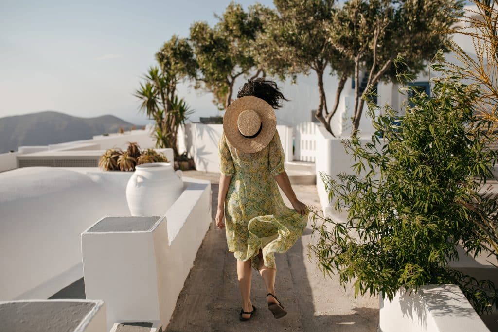 Photo of a woman from behind wearing a light green dress and a straw hat hanging around her neck. She is walking in a Greek garden.
