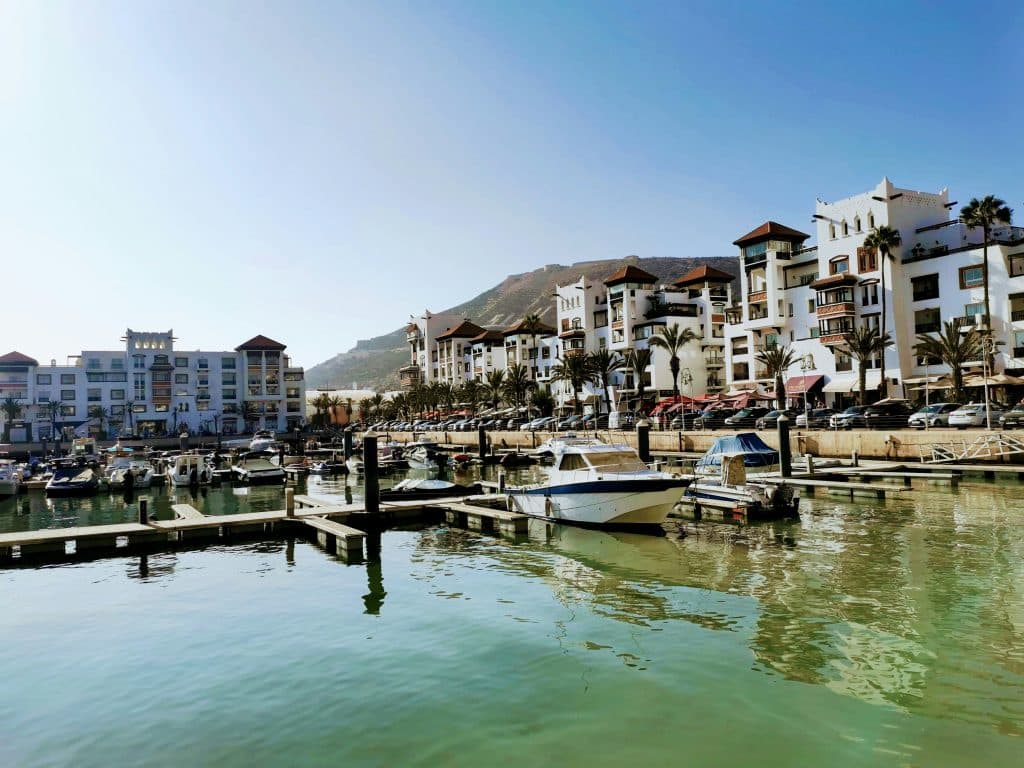 Photo of a small harbour in the city of Agadir, Morocco. A few small pleasure boats are moored in the harbour. Cars are parked opposite the harbour. The white buildings lining the harbour are well maintained and have the charming appearance of a seaside resort.