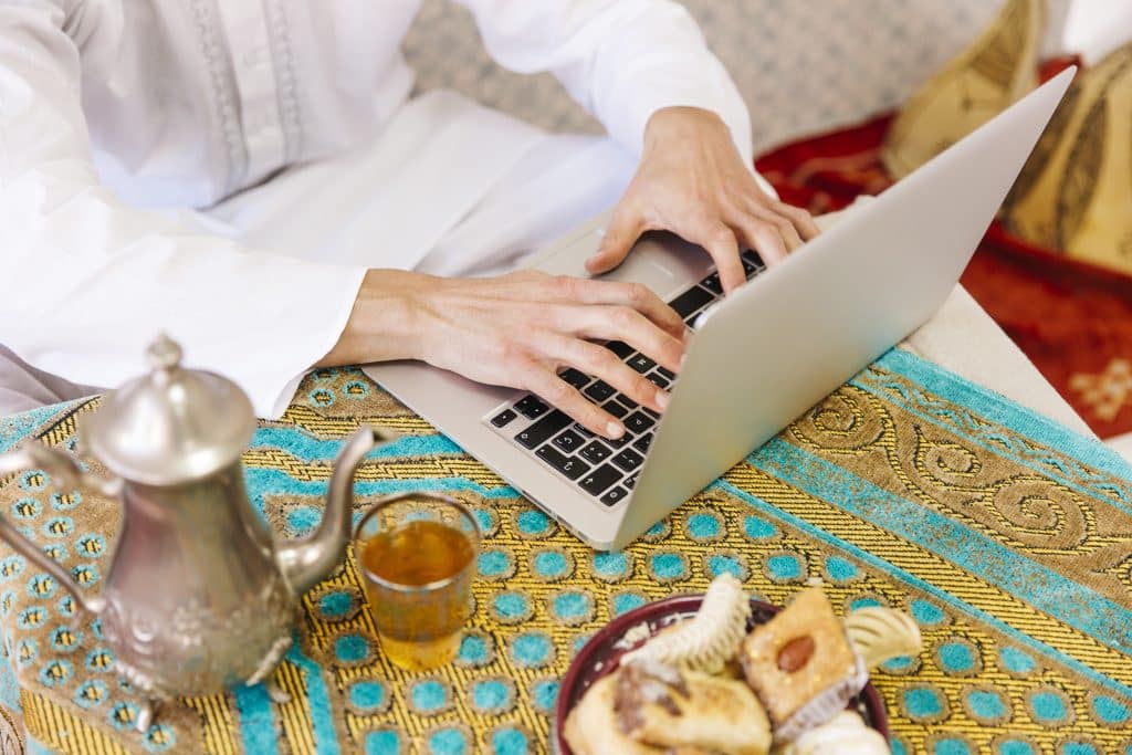 A person is working on their laptop, which is placed on a coffee table, all in a North African setting. A teapot of mint tea and sweets are placed on the table.