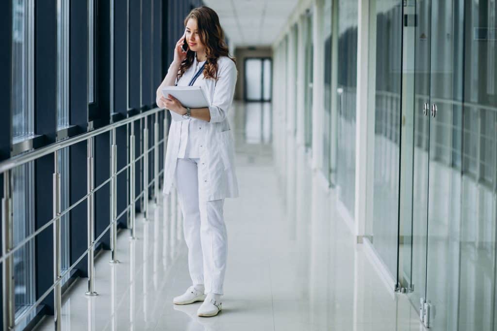 A young doctor is making a phone call in the hallway of a hospital. She is wearing a stethoscope around her neck.