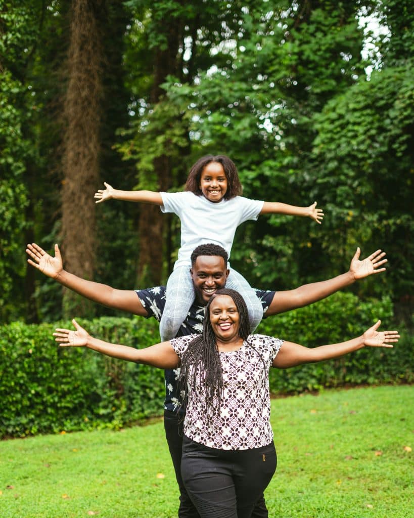 A family of three of African origin. The woman is standing in the foreground, and behind her, her husband is carrying their daughter on his shoulders. All of them are smiling broadly and opening their arms wide.