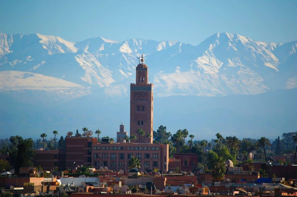 Photo of a brown concrete tower in the city of Marrakech, Morocco. The tower and its building overlook the rest of the neighbourhood. Barren mountains can be seen in the background of the photo.