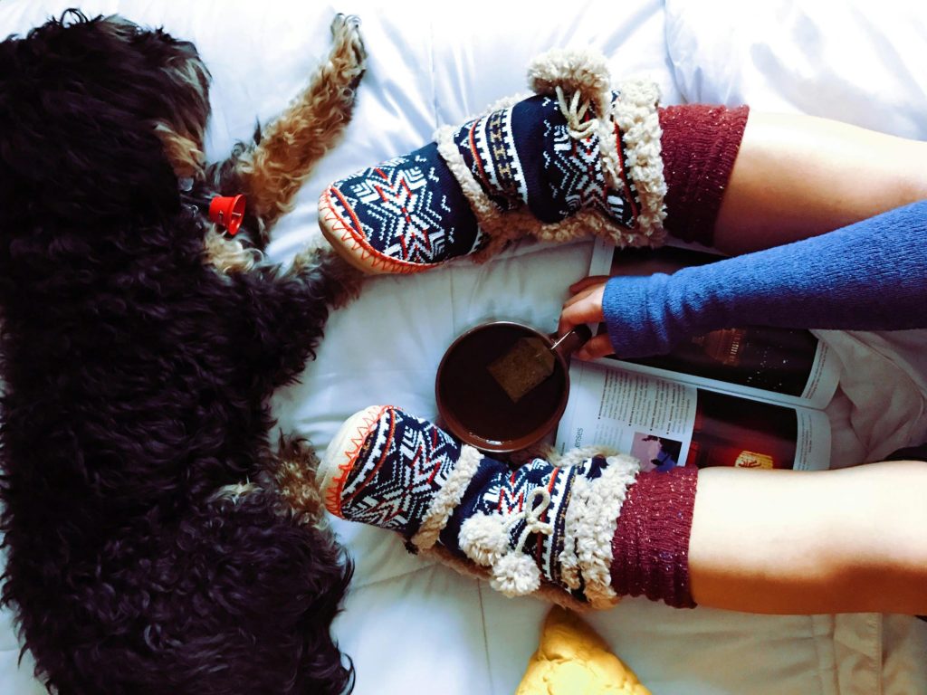 A black and beige dog is lying next to a girl wearing fluffy winter socks and mittens, who is holding a cup of tea. The dog and the girl are sitting on a bed, and the girl is reading a magazine placed between her legs.