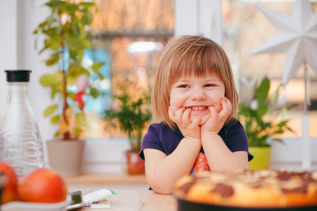 A smiling little girl holds her face in her hands, leaning on a dining table. Food and a jug of water are on the table.