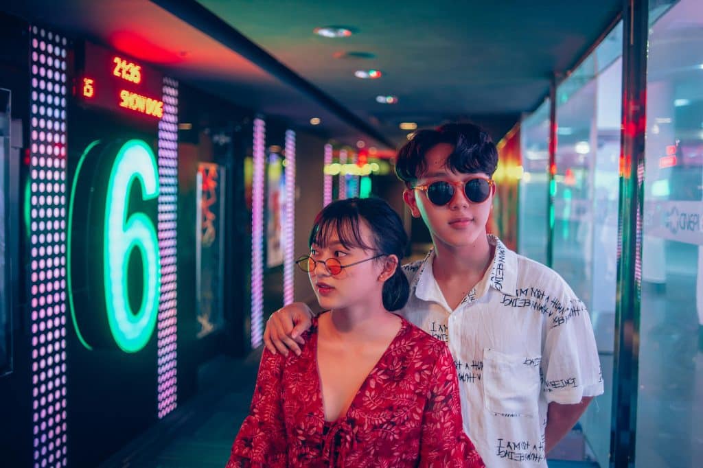 A young Vietnamese couple wearing sunglasses and dressed in a trendy style are standing in a building decorated with flashy LED lights.