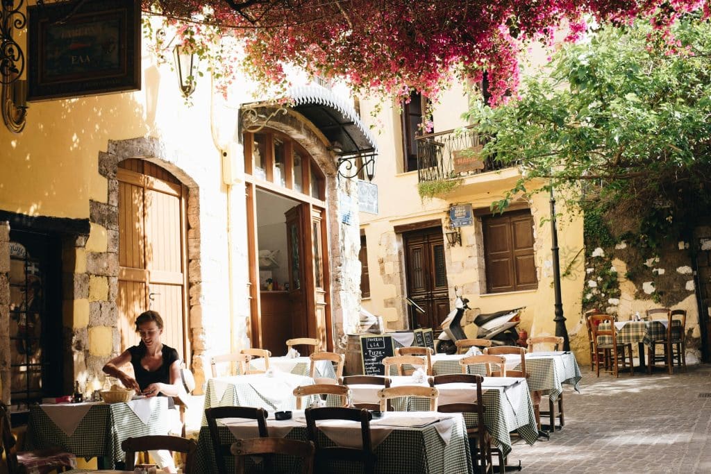 Photo of a restaurant terrace in a Greek town. The restaurant building is beige and Mediterranean in style. A woman is having lunch on the terrace.