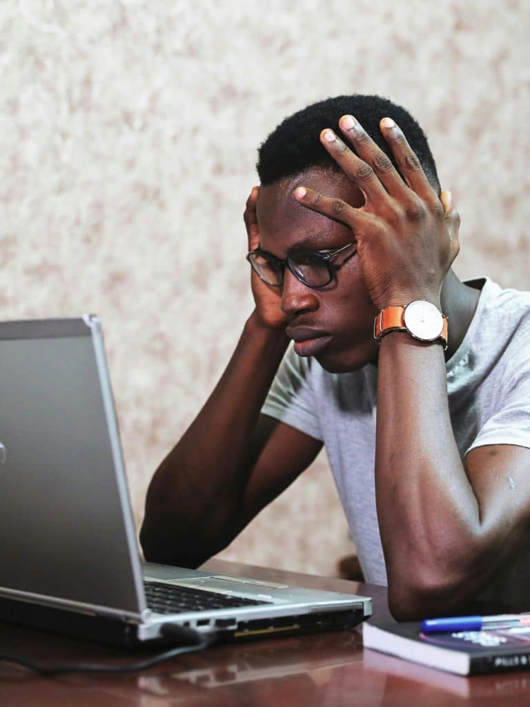 A young black man is holding his head while looking at his laptop with a worried expression.