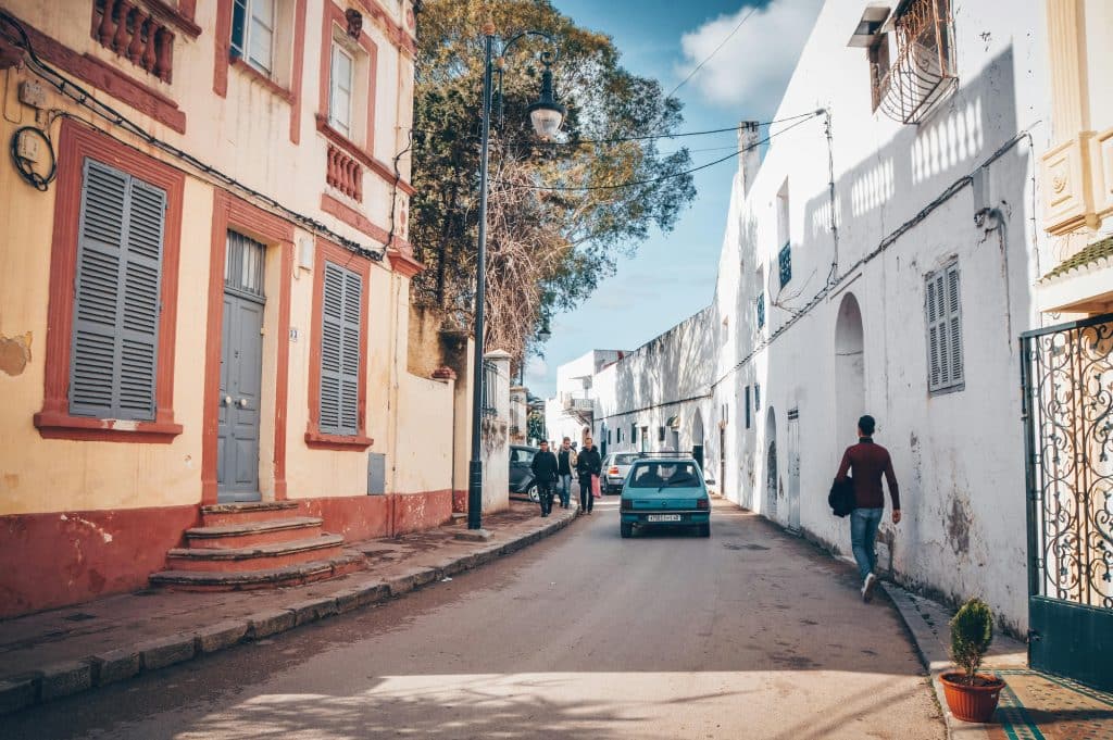 A small blue car in a narrow street in Tangier, Morocco. A few people are walking down the street, squeezing in to let the car pass.