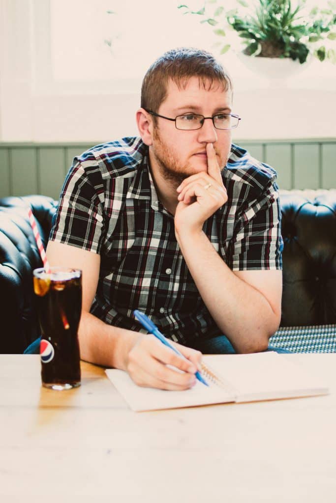 A blond man with glasses and a shirt is thinking while taking notes in a notebook. A glass of Pepsi is sitting next to his notebook. He is sitting on a black leather sofa and leaning on the coffee table.