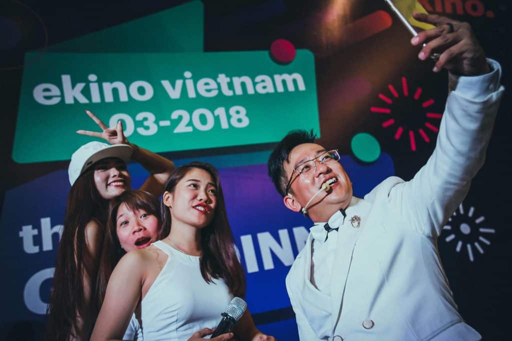 Photo taken during an event in Vietnam dated March 2018. A man in a white suit takes a selfie with three women posing for the photo, against the backdrop of a presentation screen.