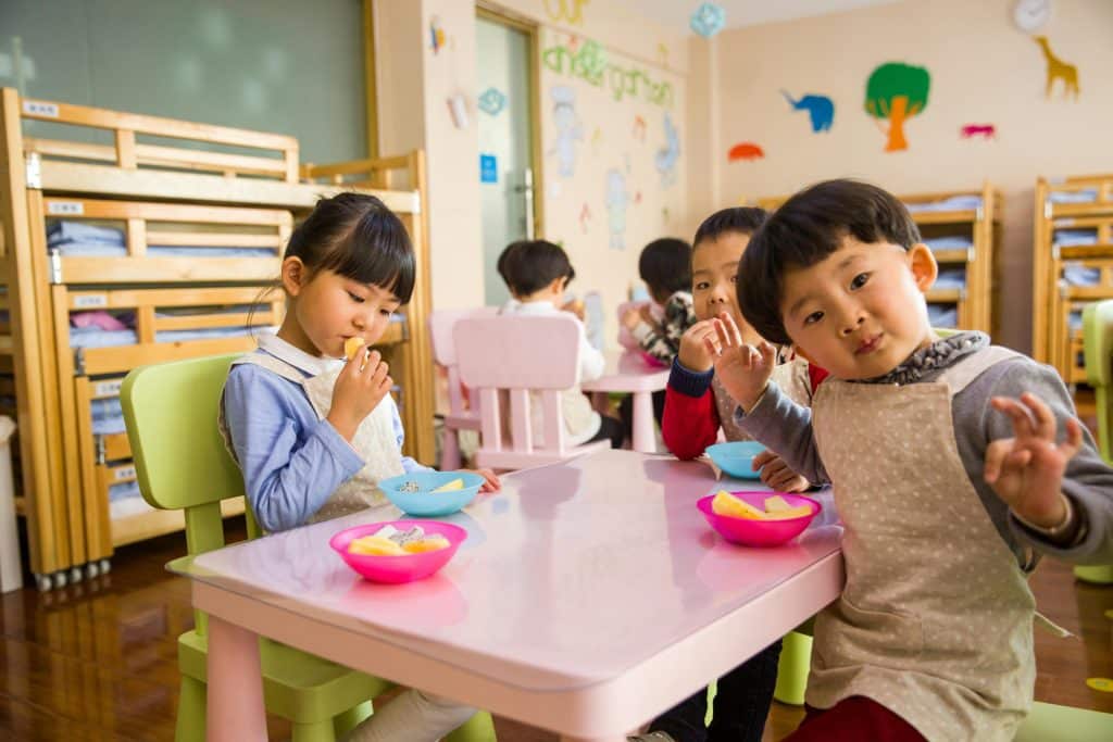 Three young children of Asian origin are sitting at a school table eating their afternoon snack. They are eating fruit and making faces at the camera.