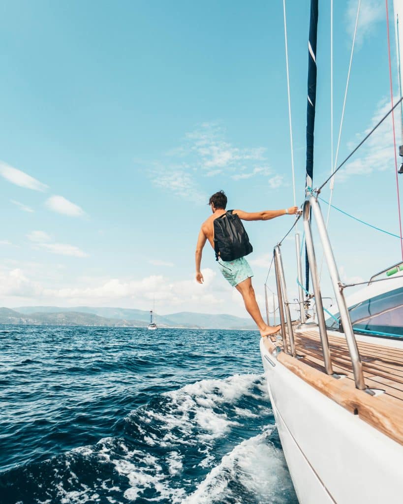 A shirtless man in swimming trunks and carrying a black backpack stands at the helm of a boat at sea and gazes at the distant horizon.