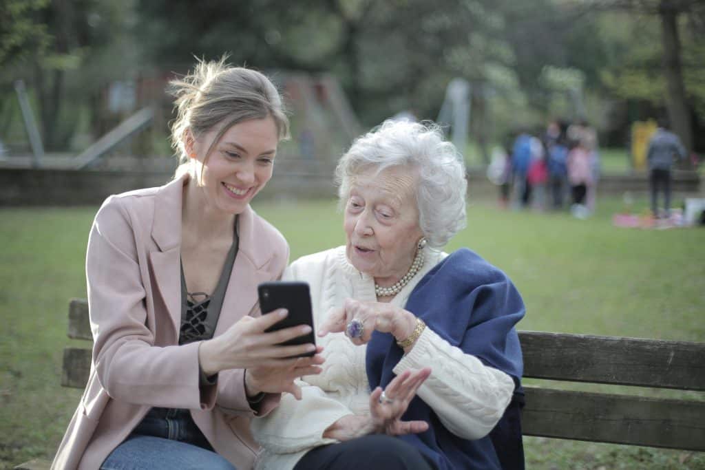 A young woman smiles as she shows something on her phone to her grandmother, both of whom are sitting on a wooden bench in a park. The young woman is wearing a light pink jacket and has her hair tied back. The elderly woman is wearing a white knitted jumper.