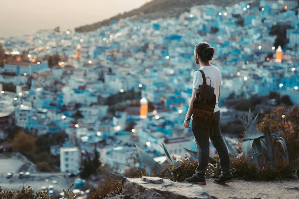 A young bearded man carrying a black backpack stands on a hill and looks out over a city in Morocco.