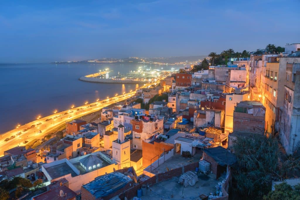 Evening photo of the city of Tangier in Morocco. The city and its streets are illuminated by streetlights and houses. The road running along the sea and the port is busy with cars.