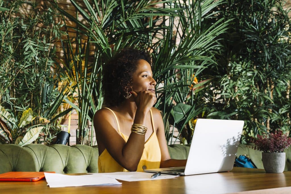 A young black woman is working on her laptop in a café with tropical décor. She smiles as she thinks. She is wearing a yellow tank top.