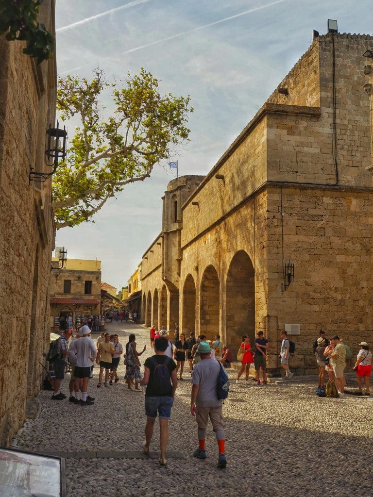 Tourists stroll through an old street in Rhodes, Greece, which is home to ancient buildings. The street is paved and has a lot of charm.