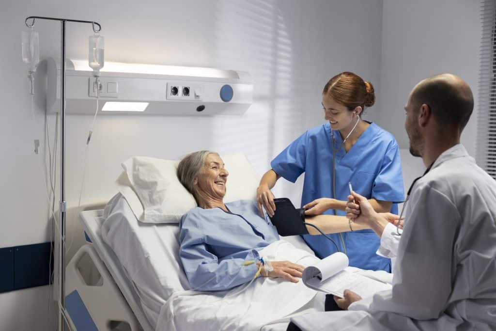 An elderly patient in her hospital bed is being examined by a nurse and a doctor. The nurse is measuring her blood pressure while the doctor talks to the patient and takes notes.
