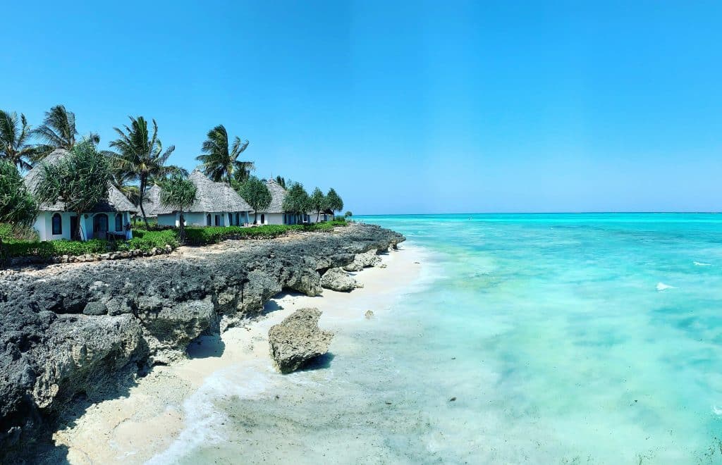 The rocky paradise beach of Zanzibar. Numerous small bungalows line the beach, surrounded by coconut and palm trees. The water is turquoise and completely transparent.