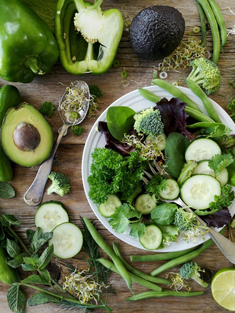Photo of green vegetables and fruit attractively arranged on a plate and on the table, with a spoon and fork. The vegetables include broccoli, avocado, cucumber, lettuce, green beans, etc.