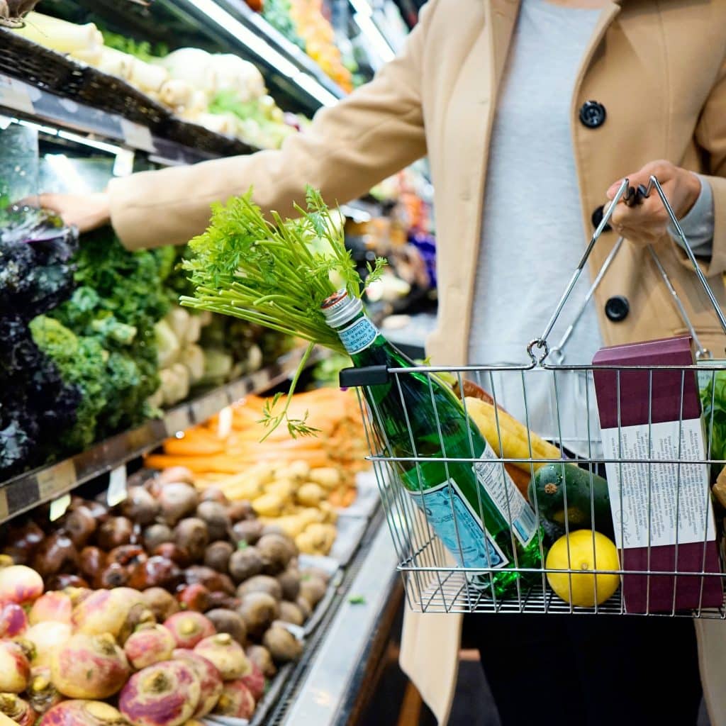 A woman in a beige coat is shopping in a store and placing her purchases in a basket, which already contains water and vegetables.