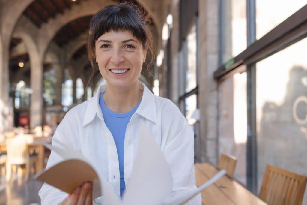Portrait of a Greek medical student in an old building with large bay windows. She is holding a notebook in her hands and smiling at the camera.