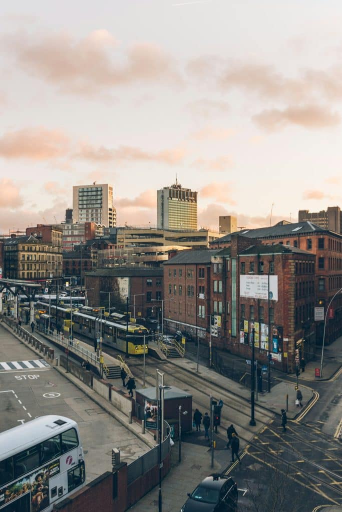 Image of a tram stop in Manchester, next to a bus station. The surrounding buildings are made of brown brick. The photo was taken in the evening at sunset. It is not rush hour as there are few people around.