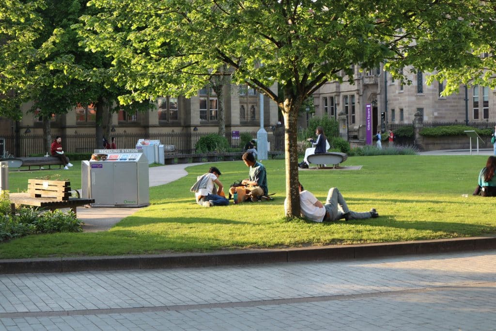 A small group of students are sitting on the grass in Gilbert Square at the University of Manchester in England. Some are sitting against a tree or on a bench.