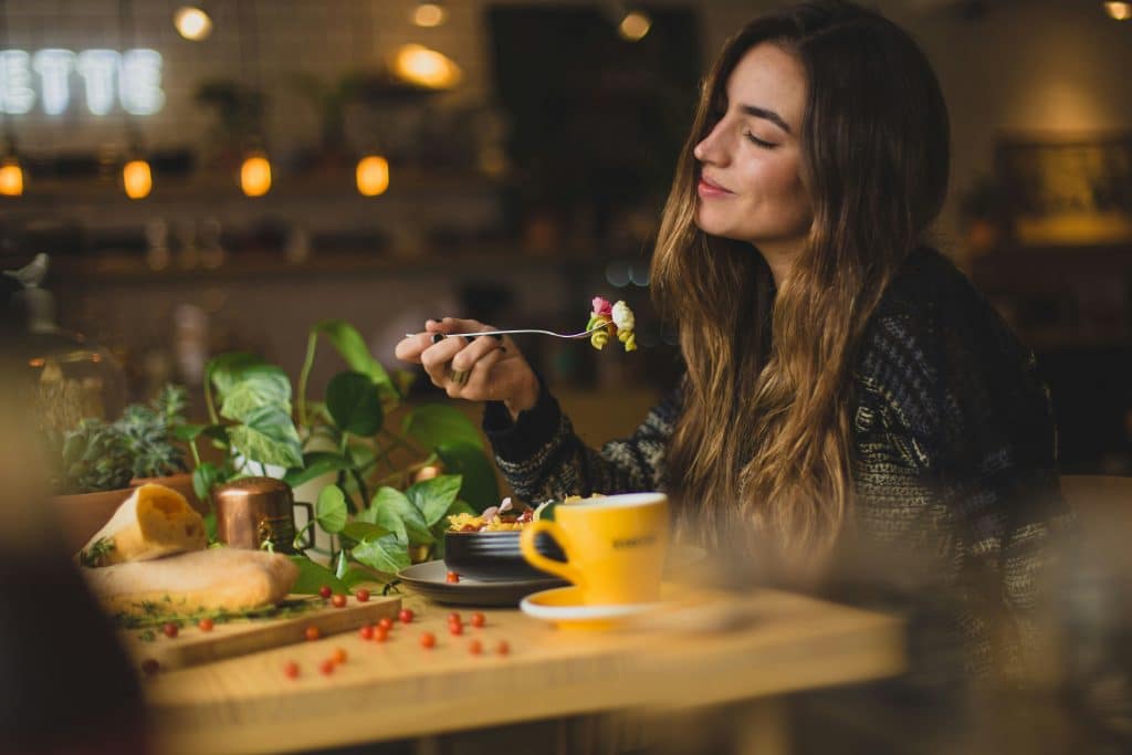 A young woman smiles as she closes her eyes to better enjoy the food she is eating with her fork. It is a pasta salad. The surrounding decor is natural.