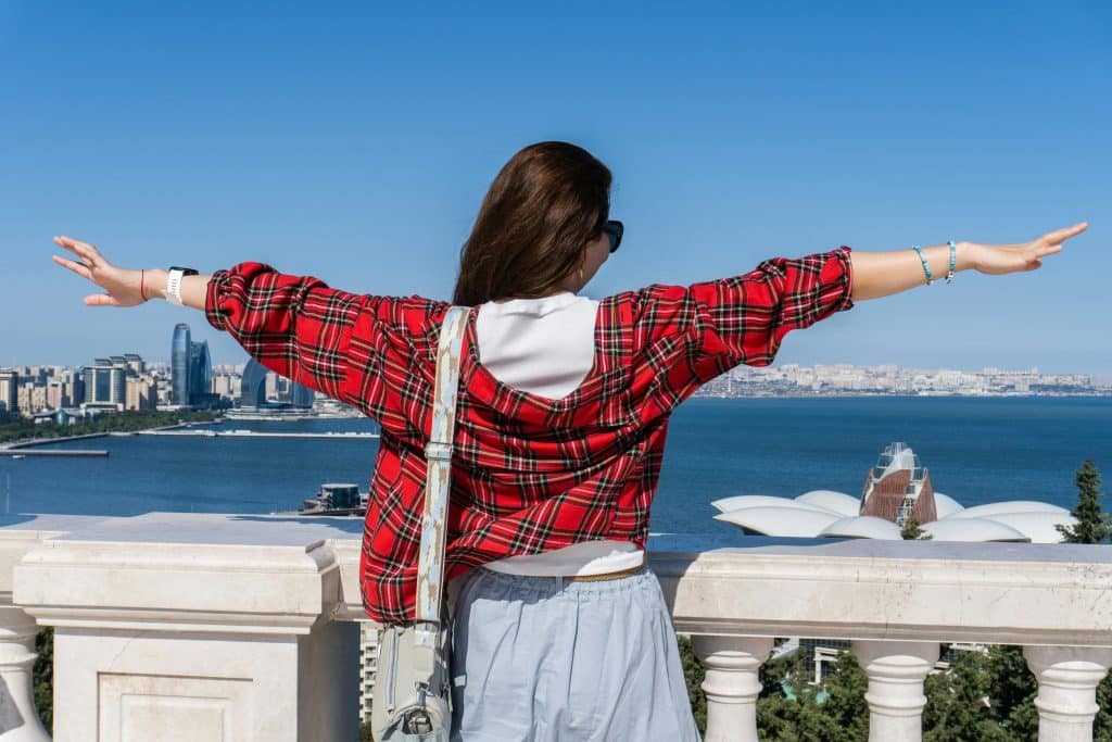A woman wearing sunglasses and a red checked shirt stands with her back to the camera on the edge of a promenade overlooking the sea in Baku, Azerbaijan. She spreads her arms out to pose for the camera.