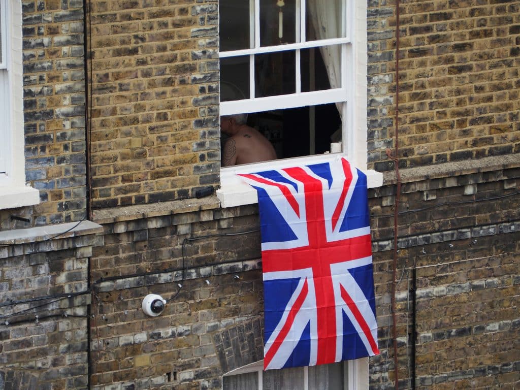 An English flag hangs from the window sill of an apartment in a building in England. A tattooed person whose face cannot be seen is sitting near the window.