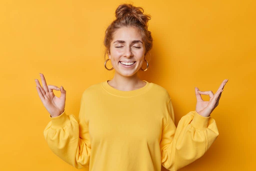 Happy calm patient young woman makes zen nirvana gesture meditates with closed eyes practices yoga has combed hair wears earrings and casual jumper smiles happily isolated over yellow background.