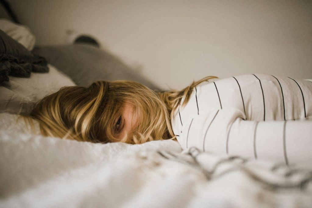 A young blonde woman wearing a striped shirt is lying face down on the bed and looking at the camera with one eye, her hair covering the rest of her face. She looks exhausted.