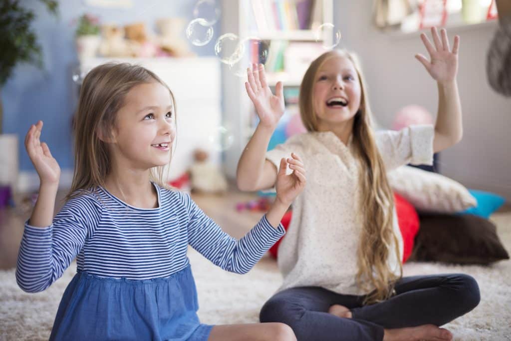 Two little blonde girls are sitting on the floor, happily playing with soap bubbles floating in the air. They are in a playroom.