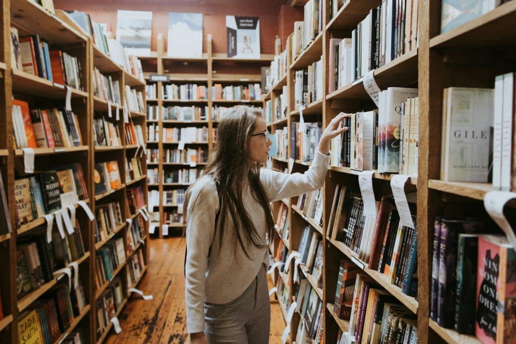 A young woman with long hair and glasses is standing in the aisles of a school library, looking for a particular book.